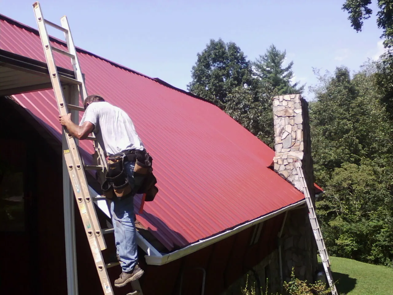 Roofer on a ladder beside a red metal roof and stone chimney — Soffit & Fascia Repair by Roofing Brothers in Pierpont